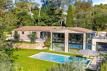 A luxurious stone house with large windows, a sparkling blue swimming pool, and lush green lawns, surrounded by abundant trees.