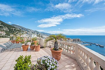 Sunny terrace with potted plants and lounge chairs, overlooking a scenic coastal bay, mountains, and blue sky.