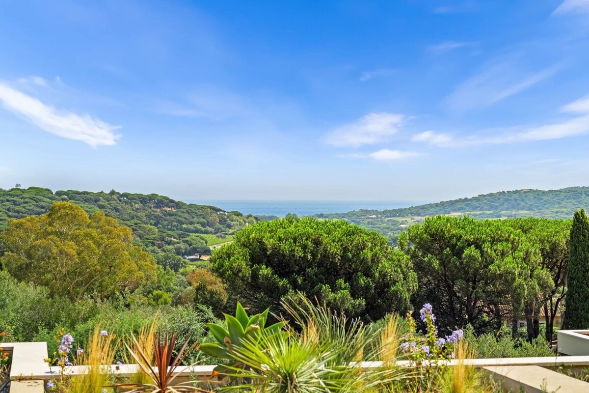 Lush green hills and trees overlook the distant blue ocean under a bright, sunny sky, viewed from a terrace garden.