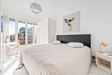 Bright white bedroom with a bed, colorful dog art, white furniture, and a large window opening to a city view.