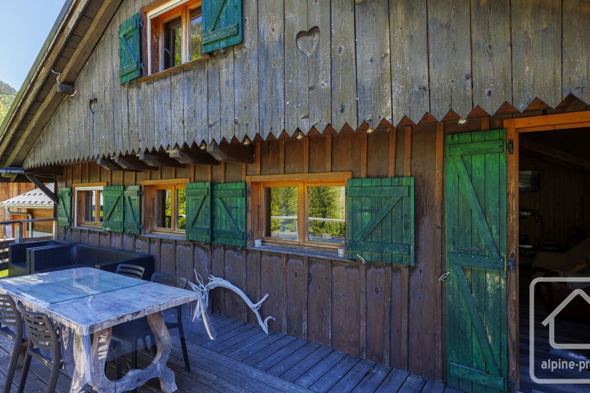 Rustic wooden chalet exterior with green shutters, a carved heart, and an outdoor dining area on its deck.