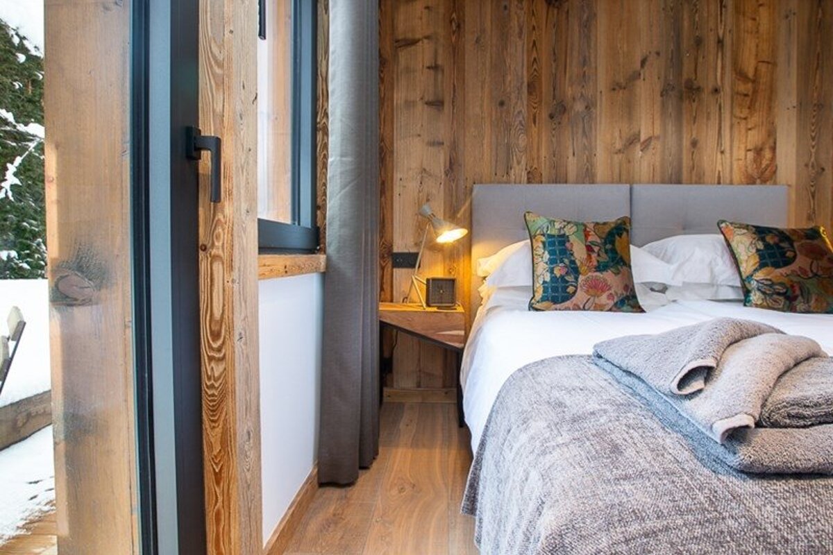 Cozy cabin bedroom with wood-paneled walls, a bed with colorful pillows, and a snowy view visible through the window.