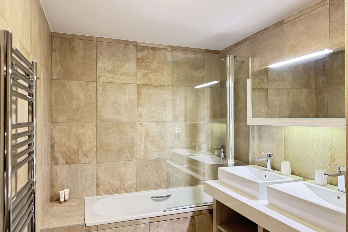 A modern bathroom with beige tiled walls, a bathtub with a glass shower screen, double vanity with rectangular sinks, and a towel warmer.