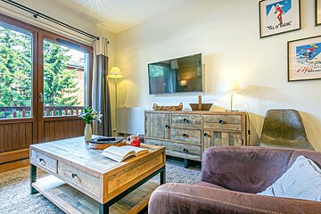 A cozy living room featuring a wooden coffee table and dresser, a wall-mounted TV, brown sofa, a balcony view of green trees, and ski posters.