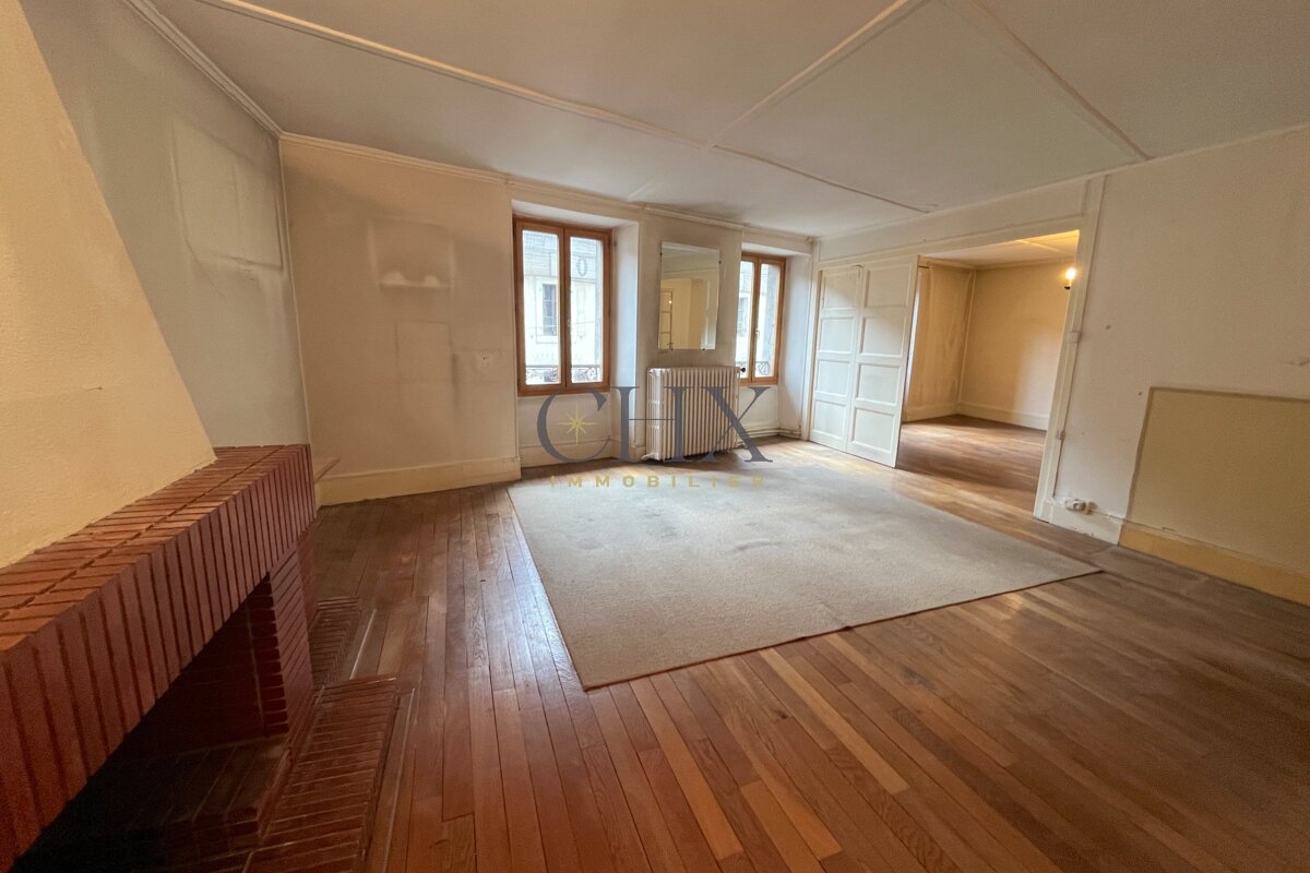 Empty room with hardwood floors, a brick fireplace, a large beige rug, two windows, and a doorway to another room.