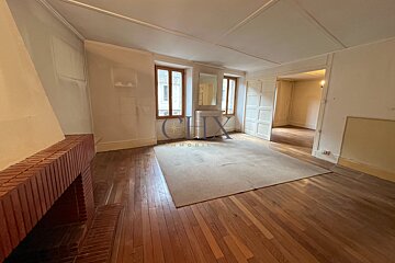 Empty room with hardwood floors, a brick fireplace, a large beige rug, two windows, and a doorway to another room.
