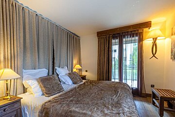A cozy bedroom featuring a large bed with a furry brown blanket, beige curtains, and warm lamps. A door/window leads to an outside view.