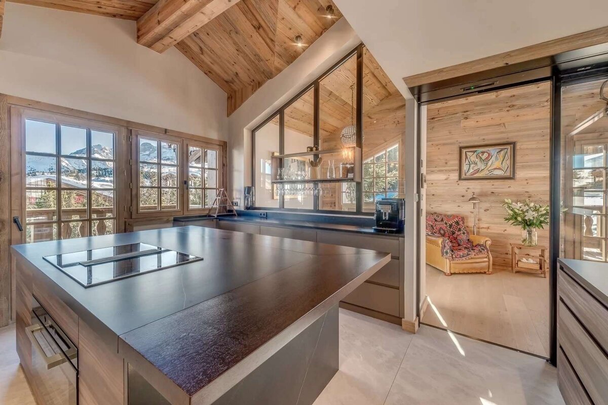 A modern kitchen with a dark island, mountain views from large windows, and wooden ceilings. Features a glass partition and an adjacent wood-paneled living room.