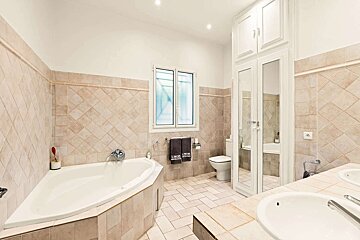 A spacious bathroom with beige tiled walls and floor, featuring a corner tub, toilet, white vanity sink, and tall mirrored storage cabinet.