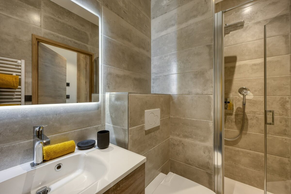 A modern, neutral-toned bathroom featuring a vanity with an illuminated mirror, a toilet, and a glass-enclosed shower, all with grey tiled walls.