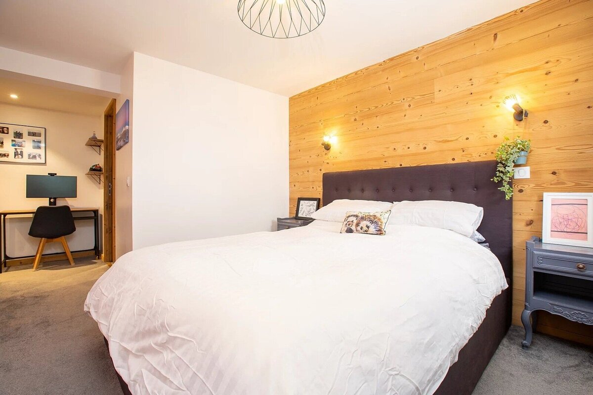 A cozy bedroom featuring a large bed with a dark headboard, a warm wood-paneled accent wall, and a desk area visible in the background.