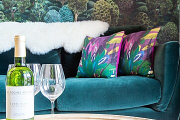 A bottle of wine and glasses sit in front of a teal velvet sofa with colorful pillows, a fluffy white throw, and forest wallpaper.