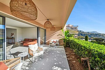A modern patio with stylish outdoor furniture, woven pendant lights, and lush greenery, offering scenic mountain and coastal views. An indoor living room is visible.