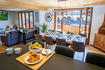 Modern chalet interior featuring a dining table set for guests, a cozy living area, and fresh pastries on a breakfast counter.