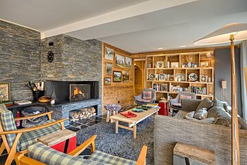 A warm, rustic chalet living room with a stone fireplace, wood-paneled walls, built-in bookshelves, and cozy plaid & gray seating.
