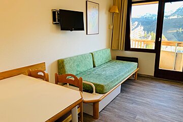 A cozy studio apartment featuring a dining table, green daybed, wall-mounted TV, and a large window overlooking a scenic mountain valley.