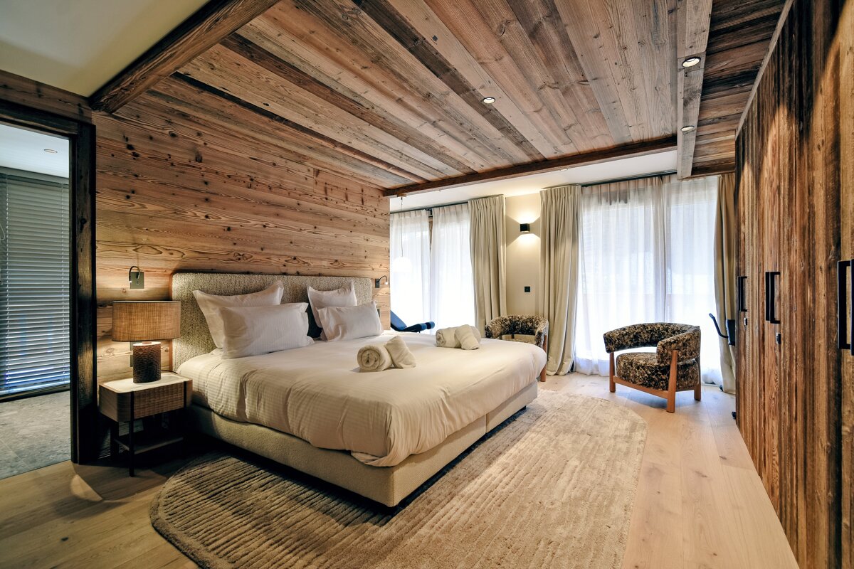 Cozy chalet-style bedroom with extensive wood paneling on walls, ceiling, and wardrobe. Features a large bed, two armchairs, and bright windows.