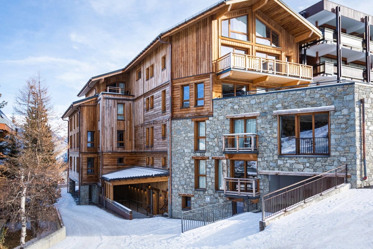 A modern chalet-style building with wooden siding and stone base sits on a snow-covered slope, surrounded by trees under a bright sky.