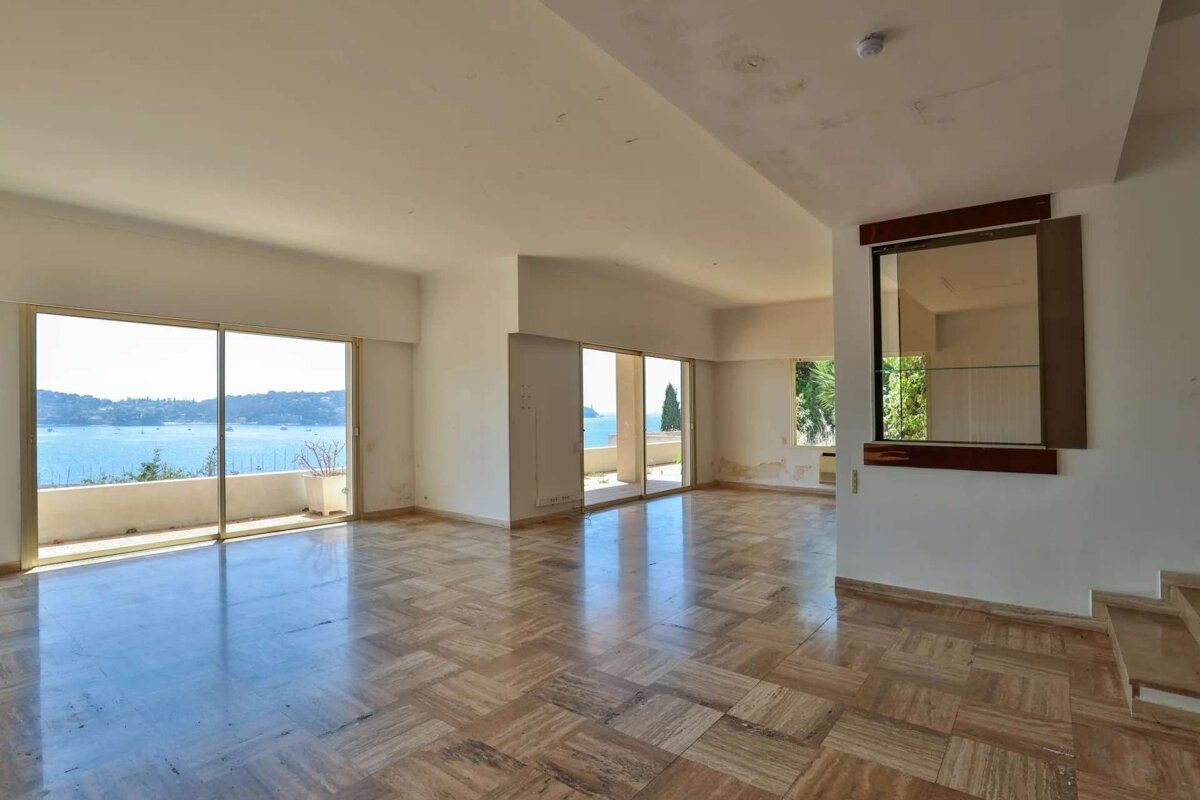 Empty, spacious room with patterned tiled floor and large sliding glass doors to a balcony, offering stunning sea views. Walls show wear.