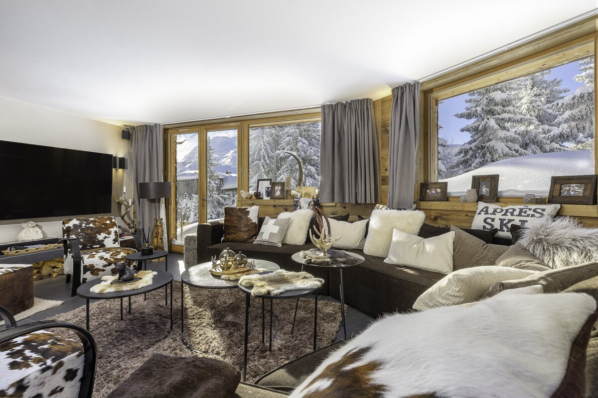 Cozy ski chalet living room with panoramic snowy mountain views. Features wood panels, dark sofa with fur, animal print seating, and decorative tables.