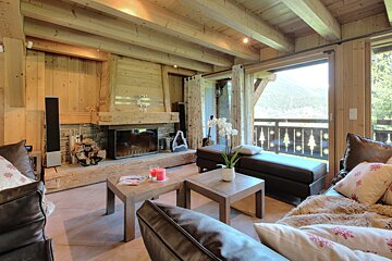 A cozy, rustic wooden chalet living room with a stone fireplace, comfortable seating, and a balcony overlooking a mountain landscape.