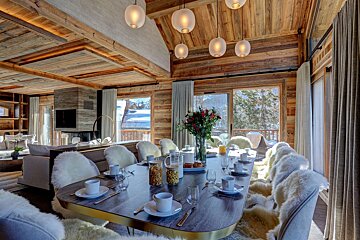 A luxurious wooden chalet dining and living area with a set table, fur-covered chairs, and large windows revealing a snowy mountain vista. Cozy fireplace nearby.