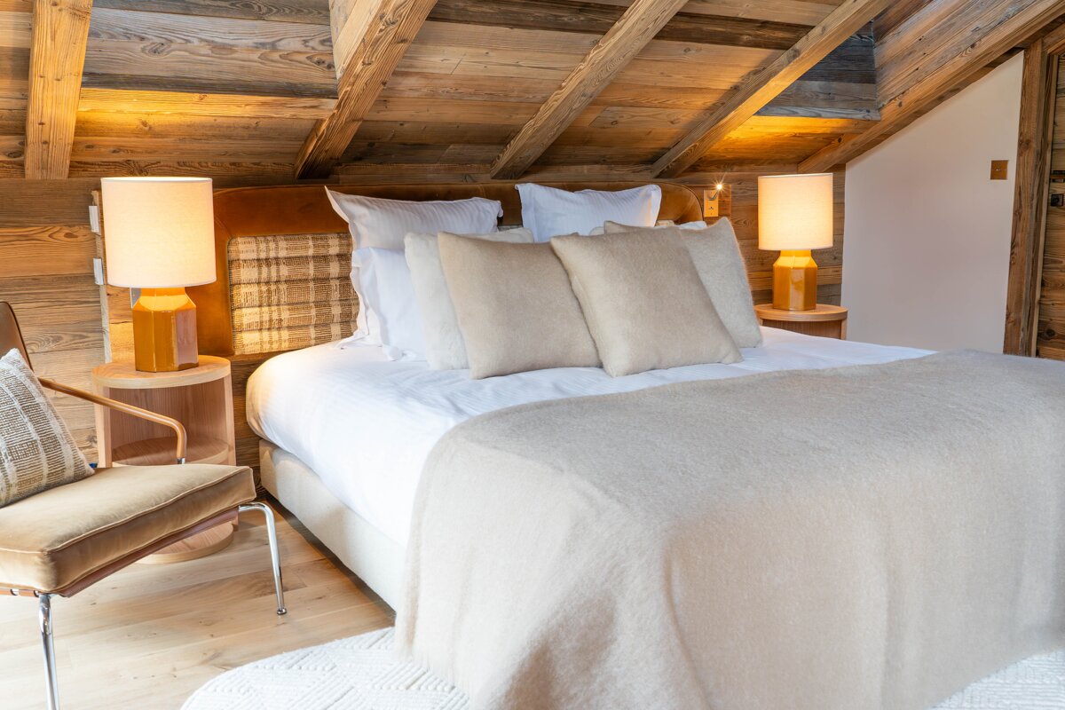 A cozy, rustic bedroom in a wooden chalet, featuring a large bed with cream bedding, two warm lamps on nightstands, and a stylish chair.