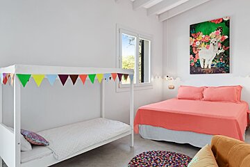 White bedroom with a canopy bed, colorful bunting, a coral bed with an alpaca painting, and a round, multicolored rug on the floor.