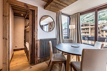 Cozy wooden chalet room with a dining table, bunk beds in an alcove, and large windows overlooking a mountain village.