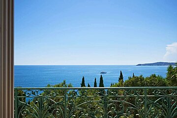 Balcony view over a clear blue sea with a yacht, green trees, and a distant coast under a bright sky.