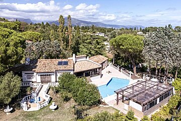 Elevated view of a villa with a tiled roof, pool, and glass-covered enclosure, set amidst lush trees and rolling mountains.
