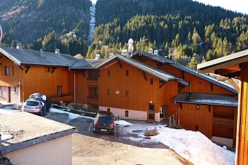 Wooden chalets in a snowy mountain resort, with pine trees and a funicular on the distant slope. Cars are parked on patchy snow.