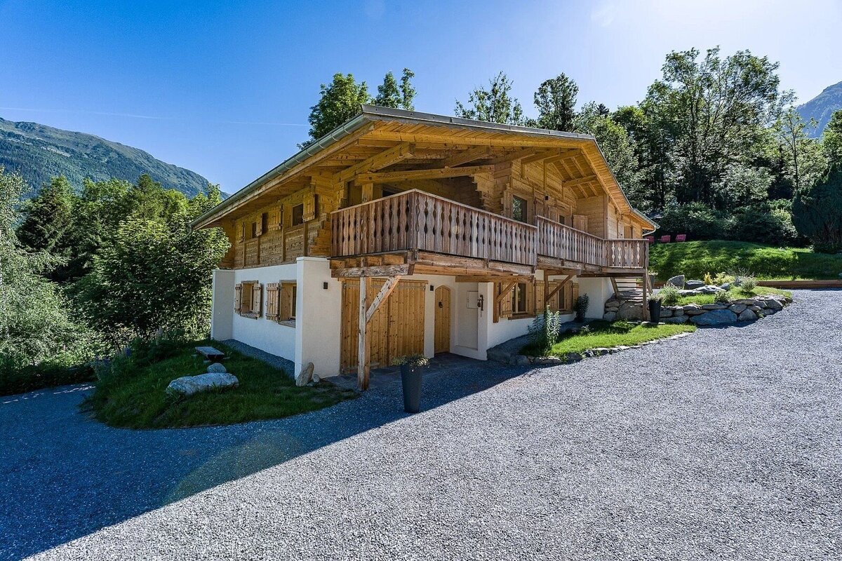 A beautiful wooden chalet with a large balcony, white ground floor, and gravel driveway set against a mountain backdrop.