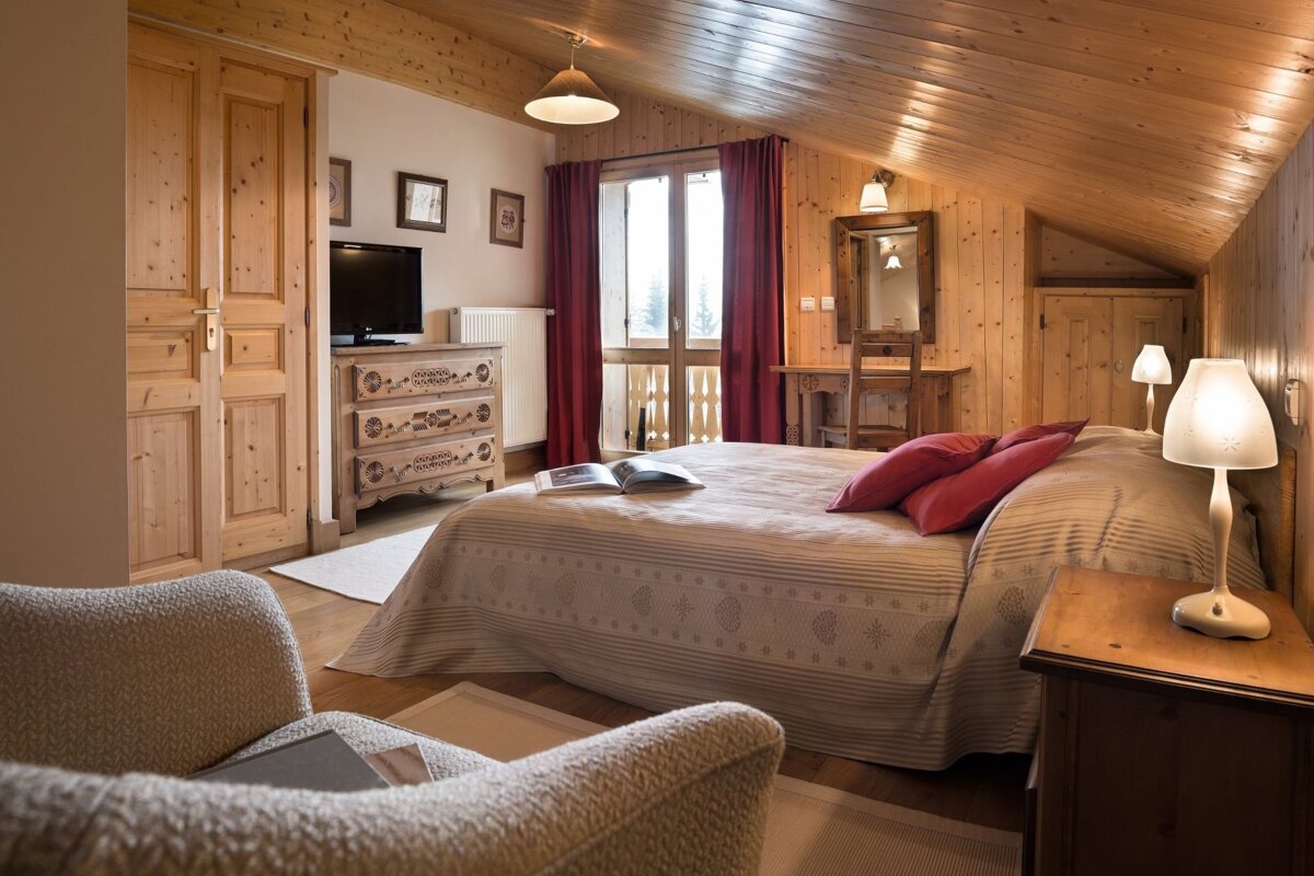 A warm, rustic bedroom with wood paneling, a large bed, red pillows, and a balcony. Features a cozy, chalet-style design.
