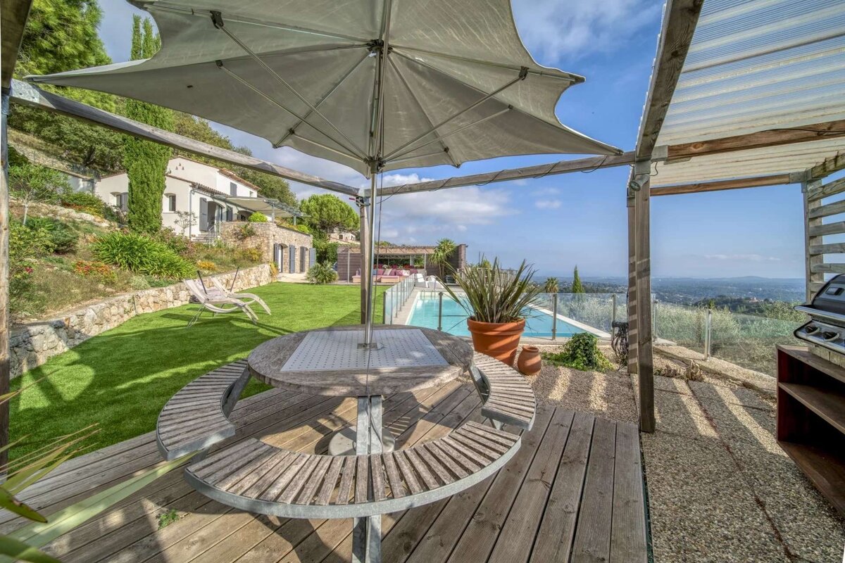 Sunny patio with a round picnic table, umbrella, and pool overlooking a green garden and hillside house. A distant scenic landscape is visible.