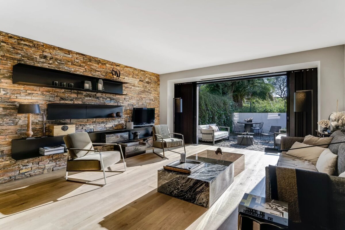 A modern living room featuring a stone accent wall, elegant furniture, and sliding glass doors that open to a lush outdoor patio.
