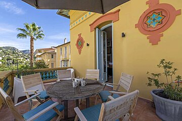 Sunny yellow balcony with outdoor seating, a palm tree, and a scenic view of a hillside town and blue sky.