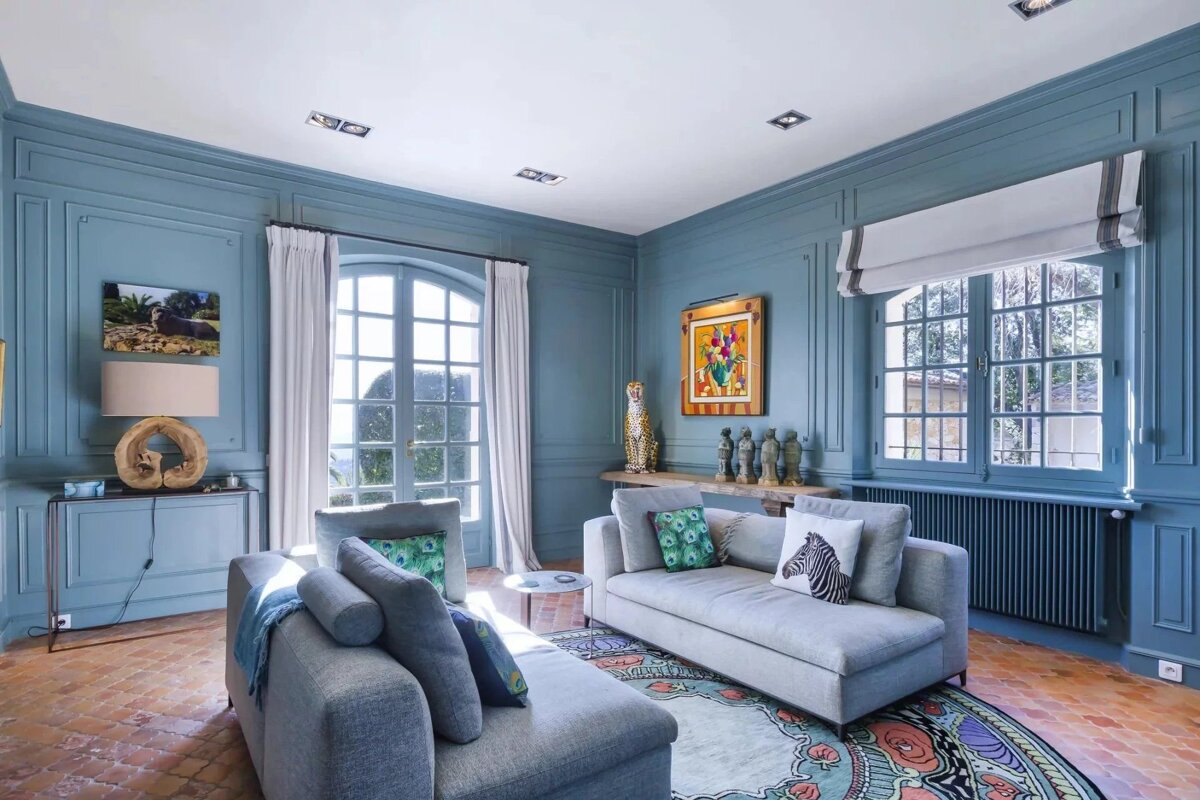 Elegant blue paneled living room with two cozy gray sofas, a vibrant round rug, colorful art, and sunlit windows creating a chic, inviting space.
