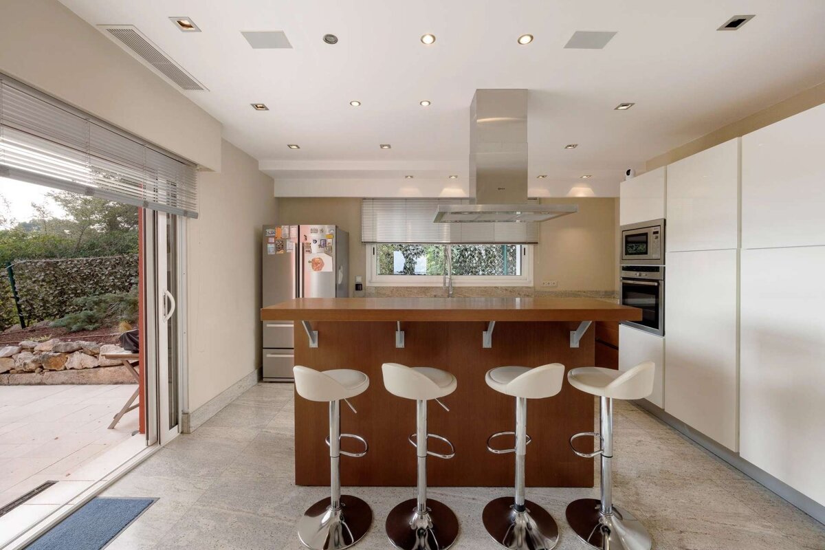 Modern kitchen with a wooden island and four white bar stools, sleek appliances, and a sliding glass door opening to an outdoor patio.