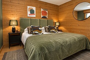 Cozy bedroom with warm wood-paneled walls, a plush green bed, abstract art prints, bedside lamps, and a round wall mirror.