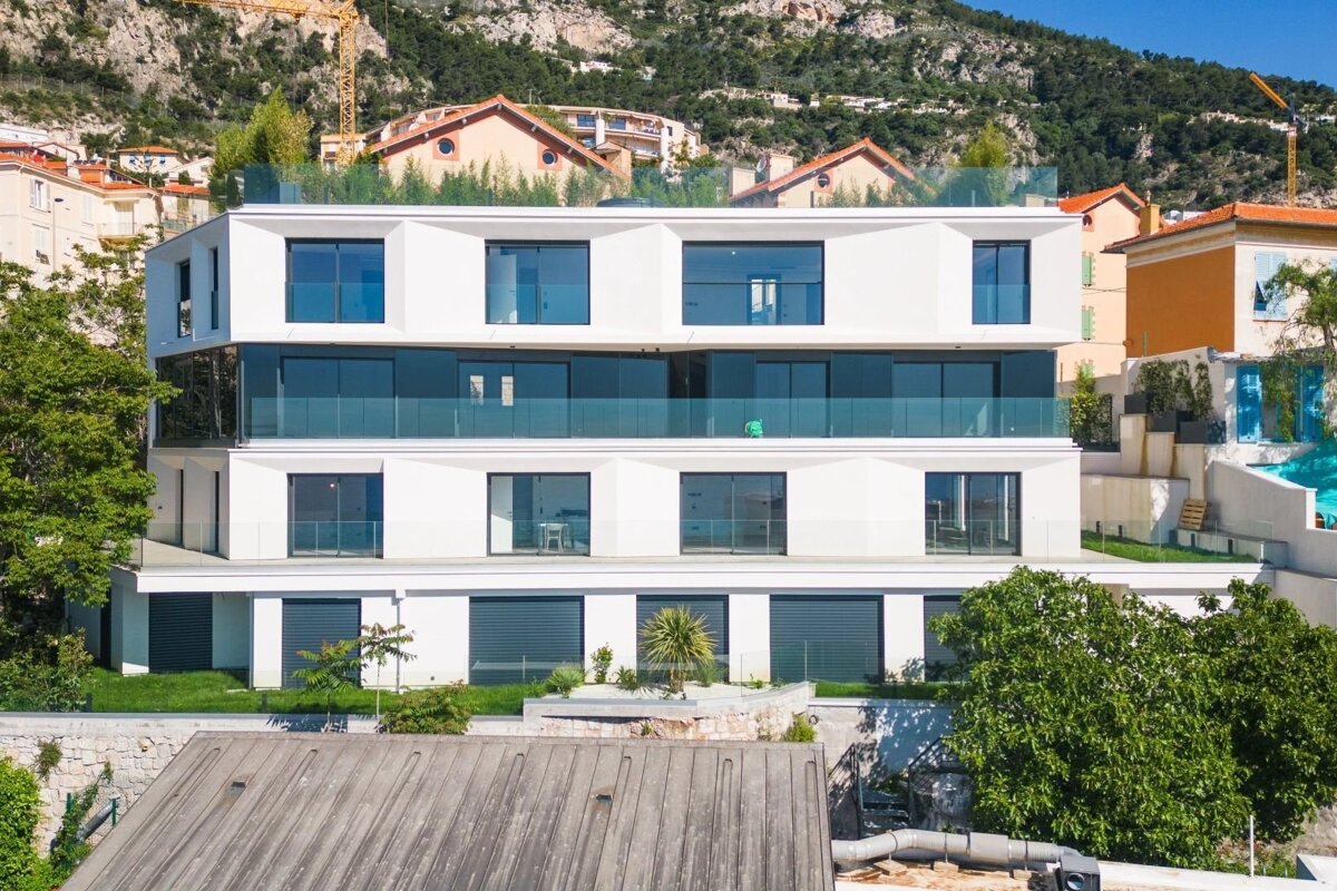 A modern, white apartment building with glass balconies, situated on a green hillside with mountains in the background.