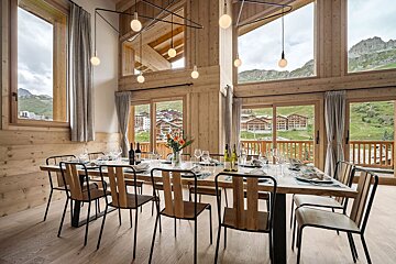 Elegant wooden chalet dining room with a long table, modern lighting, and large windows overlooking alpine scenery.