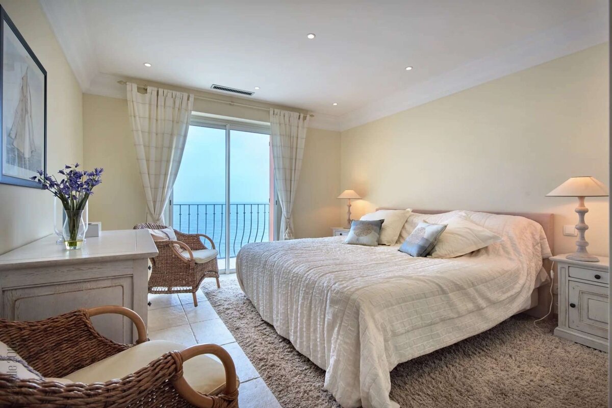 A bright bedroom with a large bed and light decor, featuring a balcony offering a stunning sea view. Wicker chairs and floral accents add to the cozy feel.