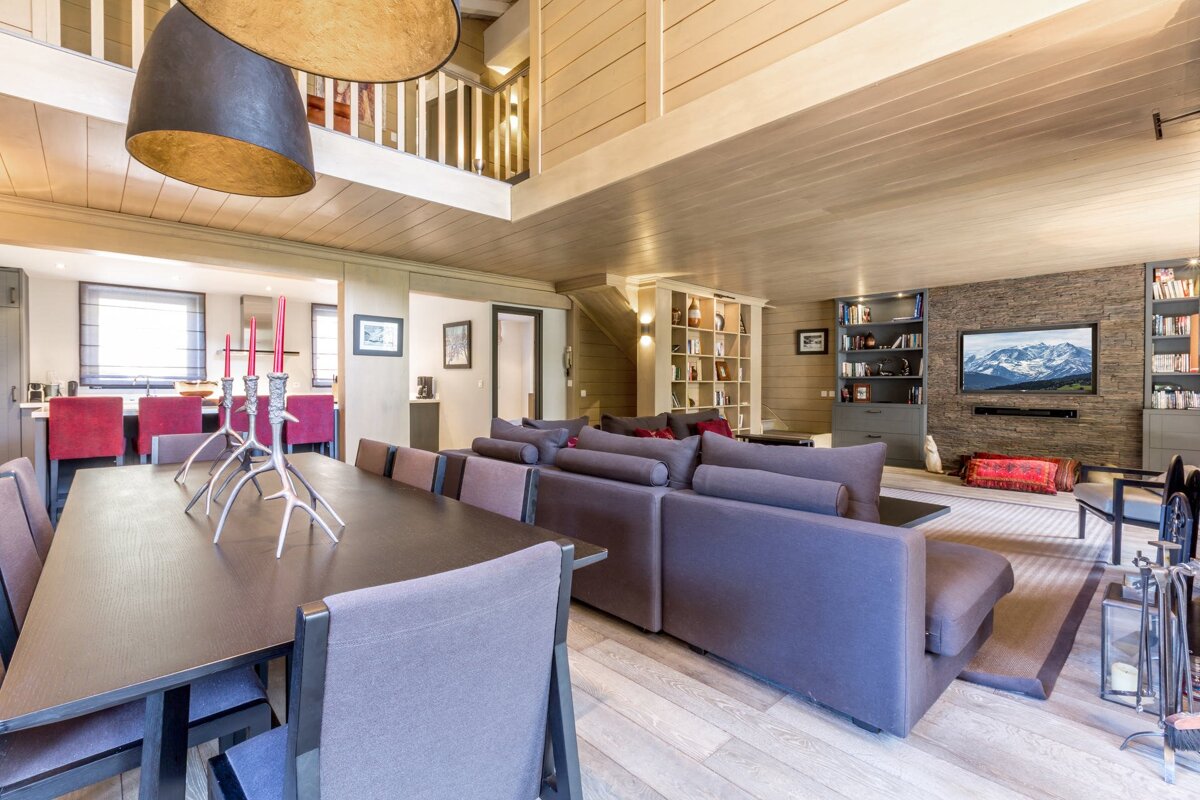 A cozy, open-plan living space featuring a dining table with unique candelabras, a large gray sectional sofa, and a TV mounted on a stone wall, with wooden finishes.