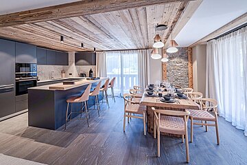 Modern open-plan kitchen and dining room with dark cabinets, wooden ceiling, and large table. Features a stone wall accent and bright windows.