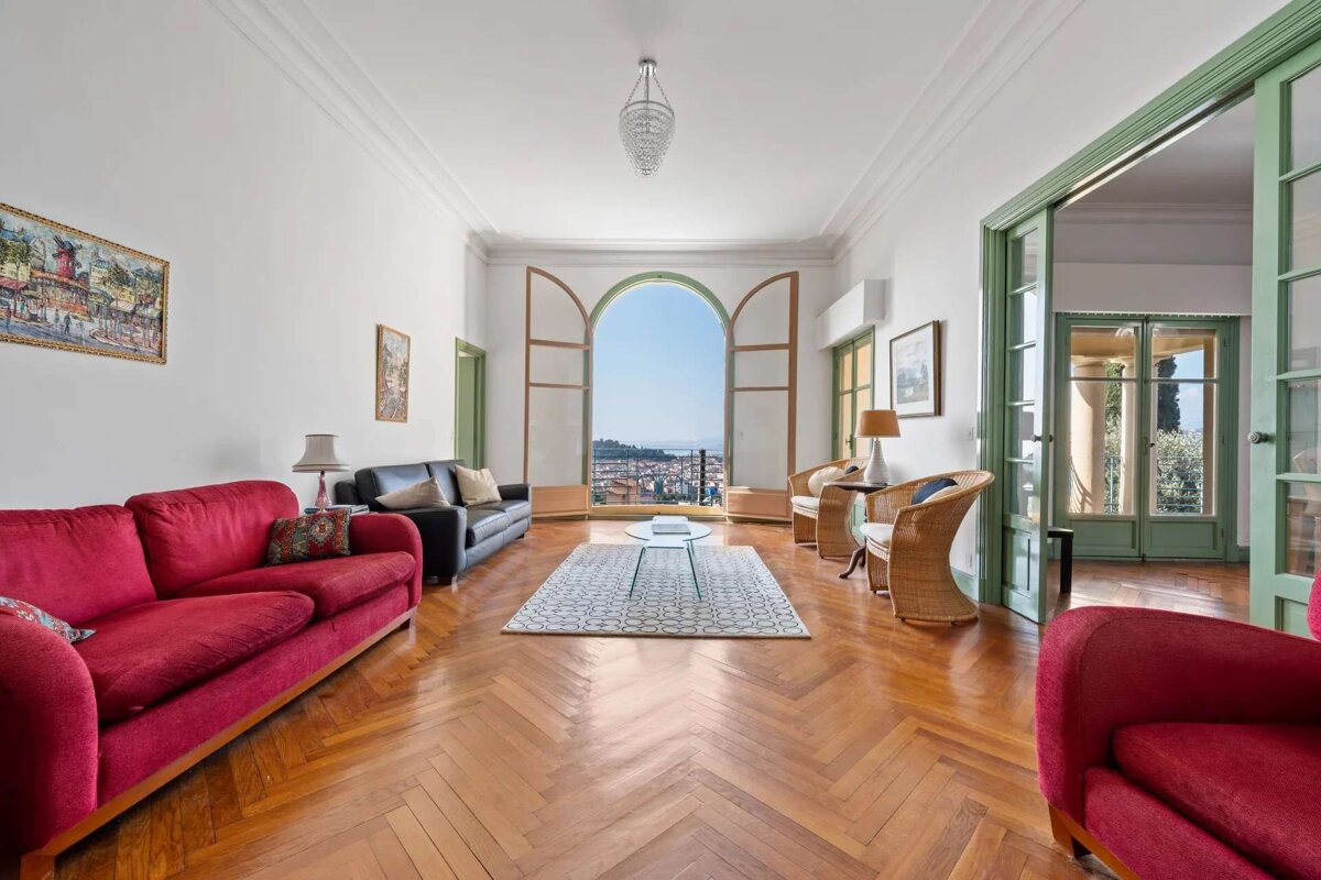 A bright, elegant living room features red and black sofas, herringbone wood floors, and a large arched window with a scenic city and sea view.