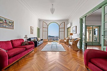 A bright, elegant living room features red and black sofas, herringbone wood floors, and a large arched window with a scenic city and sea view.