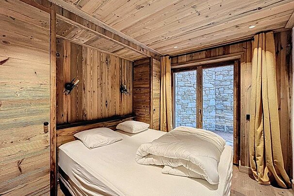 Cozy rustic bedroom with wooden walls and ceiling, a large bed with white bedding, and glass doors opening to a stone wall.