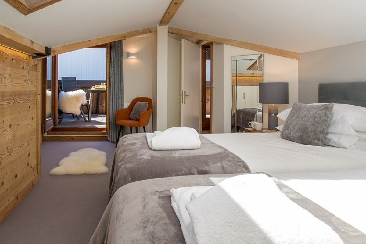 Cozy attic chalet bedroom with twin beds, an orange armchair, wooden beams, and a door leading out to a balcony.
