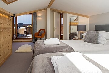 Cozy attic chalet bedroom with twin beds, an orange armchair, wooden beams, and a door leading out to a balcony.